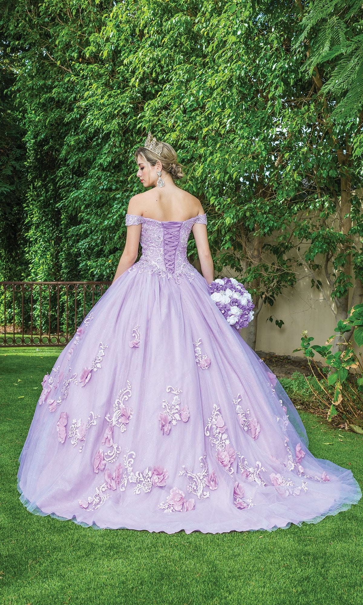 A woman wearing Quinceanera Dress 1598 By Dancing Queen, an off-the-shoulder ball gown with floral embroidery, stands on grass with her back turned. She holds a bouquet, wears a tiara, and is surrounded by lush green foliage.